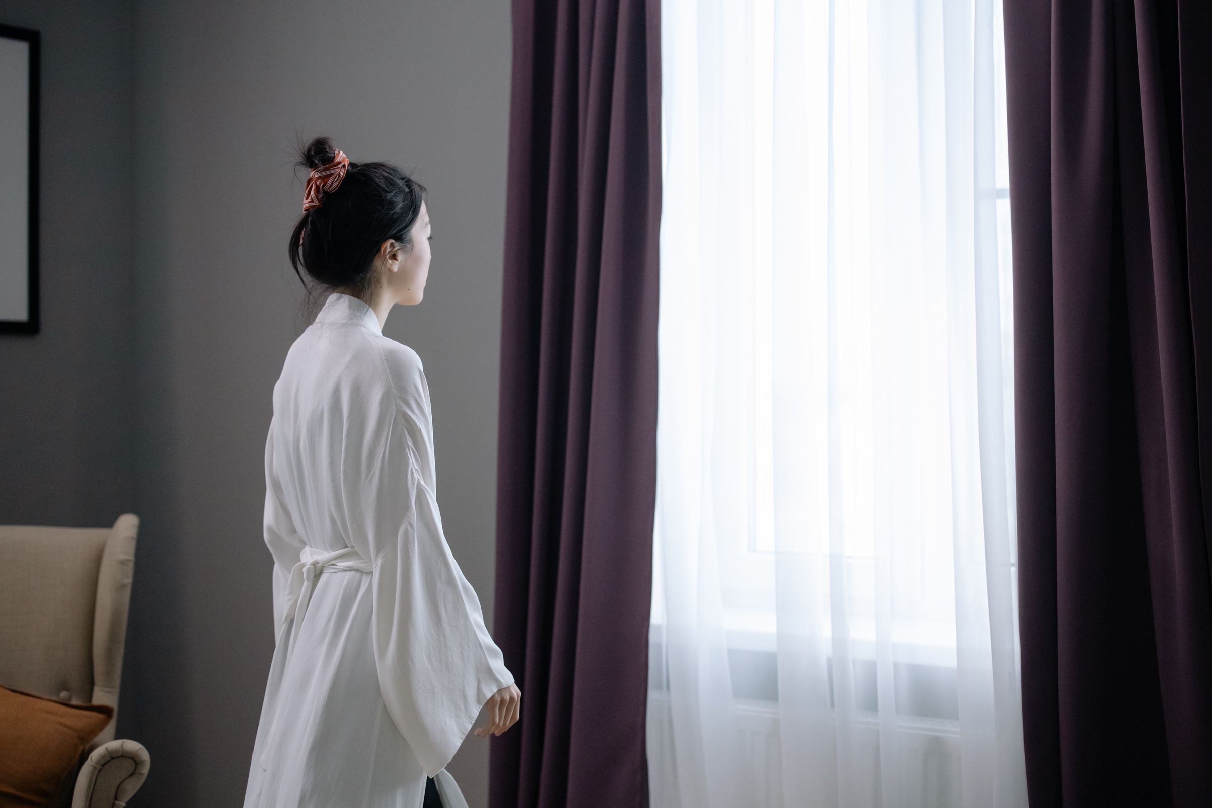 Mujer en bata blanca de pie junto a una ventana con cortinas, luz de mañana