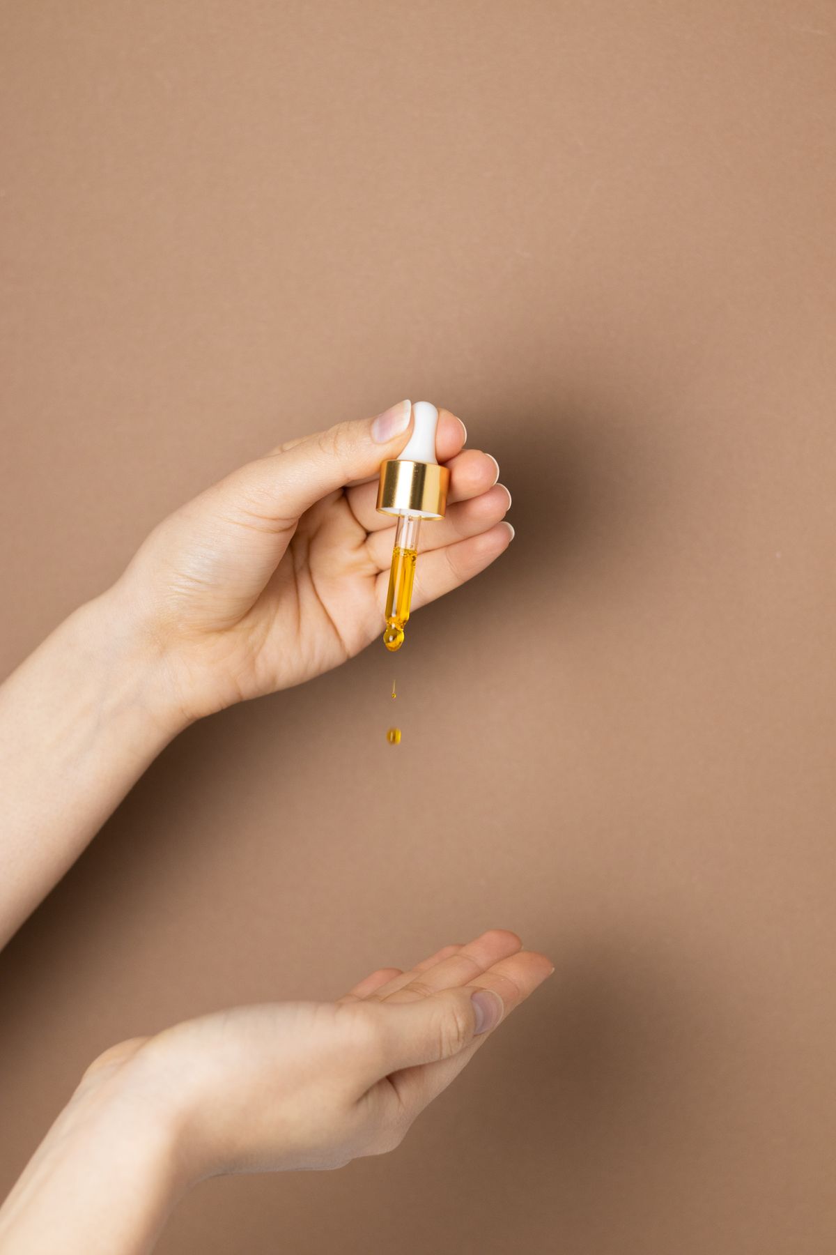 Una mano deja caer una gota de aceite desde un gotero, sobre fondo color arcilla